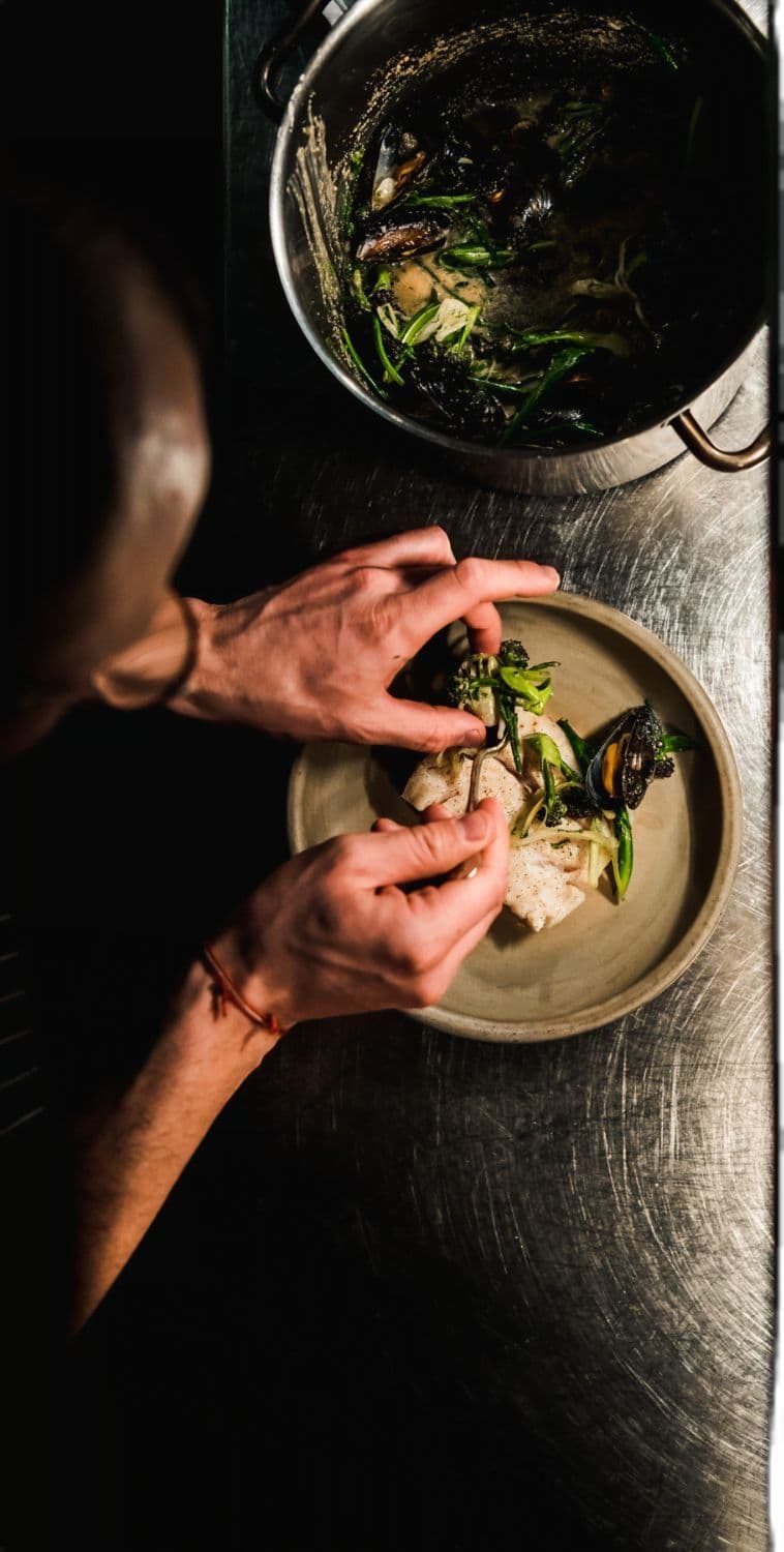 Chef carefully plating a delicate seafood dish with greens over a steel bench