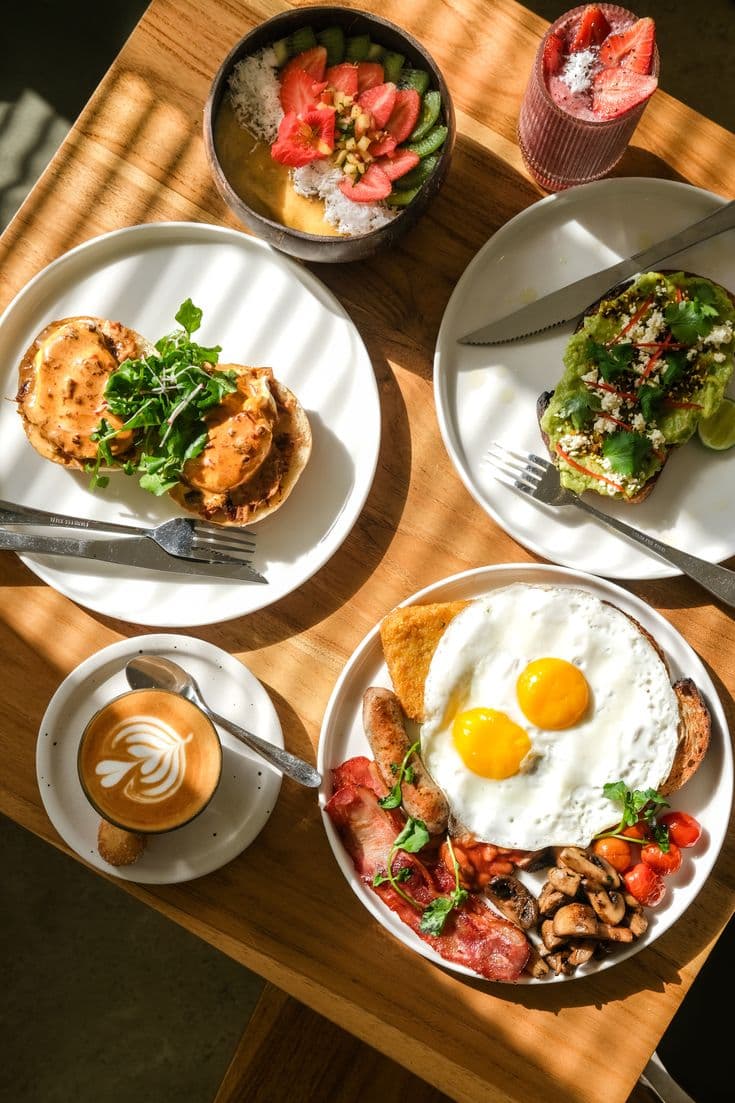 Sunlit brunch spread with a full breakfast plate, avocado toast, smoothie bowl and a latte